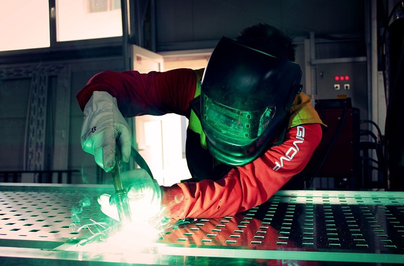 Taber Extrusions Social Study: Aluminum’s Economic Impact An image of a person wearing a black helmet and a thick red jacket welding a piece of metal, with green light sparking out of the welding machinery.