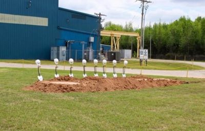 Angle on 8 new shovels with white Taber Extrusions hard hats resting neatly on each shovel handle, resting on holders just beyond a mound of freshly turned dirt from a nicely manicured lawn area in front of Taber’s Russellville, Arkansas aluminum extrusion facility.
