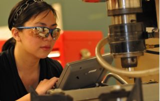 A photograph featuring a person with long hair in a ponytail held down by a headband that looks like a chain and wearing blue rimmed protection glasses that reflect the room, looks at a panel carefully. Behind the panel is a friction stir welding spinner, which looks like a big hydraulic cylinder with a drill bit at the end and there is a plastic hose attached. Behind this, a red, metallic workbox and green wall is slightly out of focus. Must be read in the voice of the movie Voice-Over artist: “In a world where dissimilar metals cannot be joined, one path stands above the rest…”