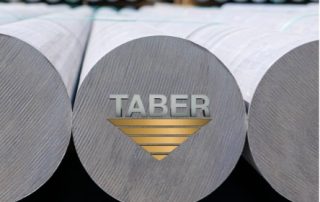 Close-up shot of the Taber logo, the letters Taber in “metal” gray and triangular metal gold logo beneath, centered and overlayed on the end of center round aluminum billets in the foreground, several more rounds blurred in the background.