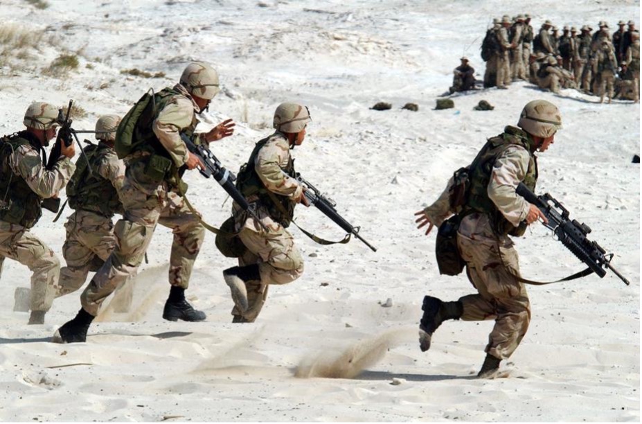 Modern day American soldiers carrying rifles run through thick sand, while beyond a large group of soldiers stand in a tight group.