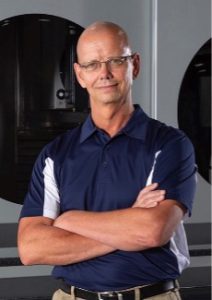 Charles Stout, President of Taber Extrusions wears a blue and while golf shirt, tan khaki pants and a black belt with his arms folded neatly across his front as he kindly smiles for the camera.
