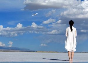 A woman in a white dress stands on the beach gazing into the summer sky at an airplane ascending into the clouds.