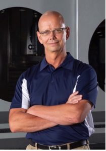 Charles Stout, President of Taber Extrusions wears a blue and while golf shirt, tan khaki pants and a black belt with his arms folded neatly across his front as he kindly smiles for the camera.
