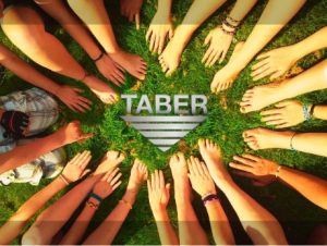 A circle of hands and feet from a group of people on luscious green grass with the white version of the Taber Extrusions logo floating in the middle.