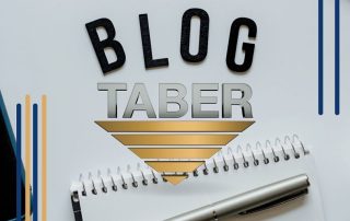 A black paper cutout of the word “BLOG” stands out, with a notebook and pen beneath. In the center, the Taber logo is encircled by L-shaped borders in gold and blue, creating a stylish composition.