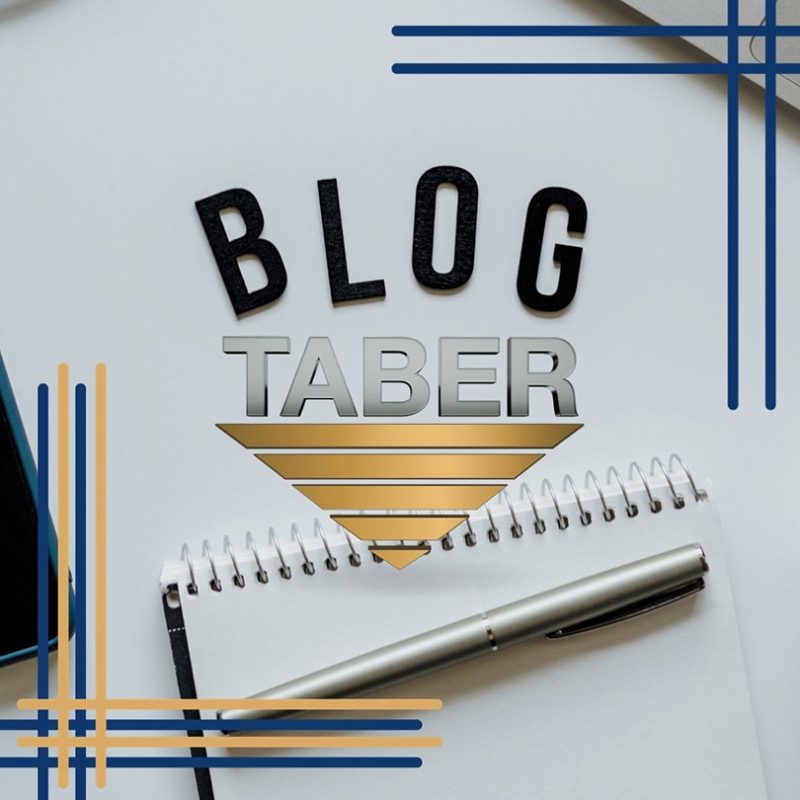 A black paper cutout of the word “BLOG” stands out, with a notebook and pen beneath. In the center, the Taber logo is encircled by L-shaped borders in gold and blue, creating a stylish composition.