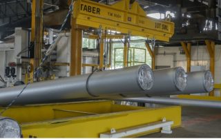 The most noticeable object in the photo is a massive yellow crane with the name “TABER” on it. The crane is lifting 3 lengthy metallic pipes that measure approximately two feet in diameter. Straps securely attach the pipes to the crane. Other industrial equipment and machinery fill the background of the image. The photo diffuses the lighting, creating a soft glow. At the same time, muted colors add to the overall industrial feel of the scene.
