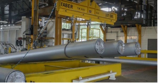 The most noticeable object in the photo is a massive yellow crane with the name “TABER” on it. The crane is lifting 3 lengthy metallic pipes that measure approximately two feet in diameter. Straps securely attach the pipes to the crane. Other industrial equipment and machinery fill the background of the image. The photo diffuses the lighting, creating a soft glow. At the same time, muted colors add to the overall industrial feel of the scene.