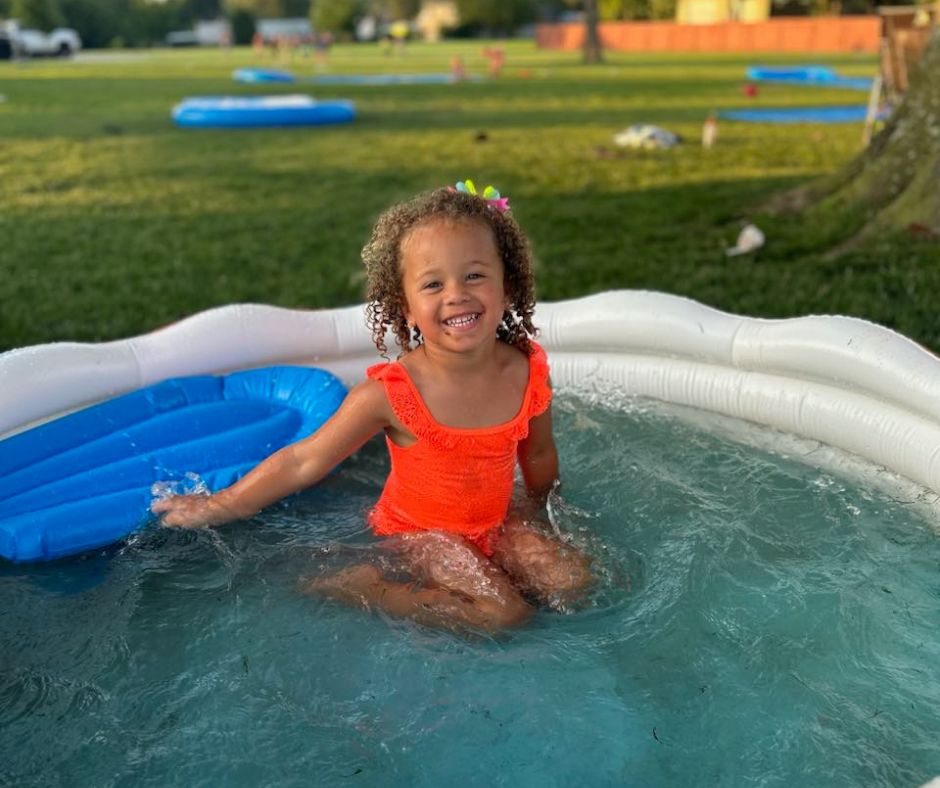 An adorable photo of JP's youngest, Savvy, smiling in her orange swimsuit sitting in an inflatable pool.
