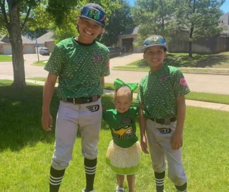 Photo of JP's smilimg three kids, the two boys wearing green baseball uniforms and their little sister between them, rocking a green t-shirt, skirt, and bow in her hair.