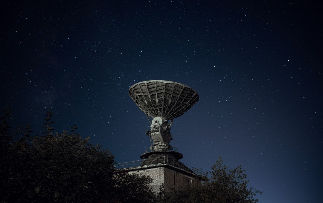 Large satellite dish under a starry night sky, with surrounding treetops in the foreground.