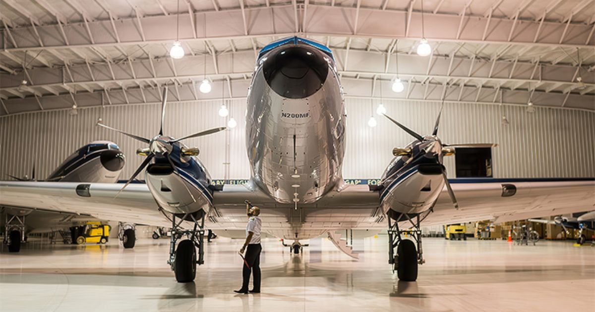 Taber-2026-08-plane-inspection-in-hangar A polished aircraft constructed with aluminum extrusions positioned inside a large hangar, with an inspector examining the structure.